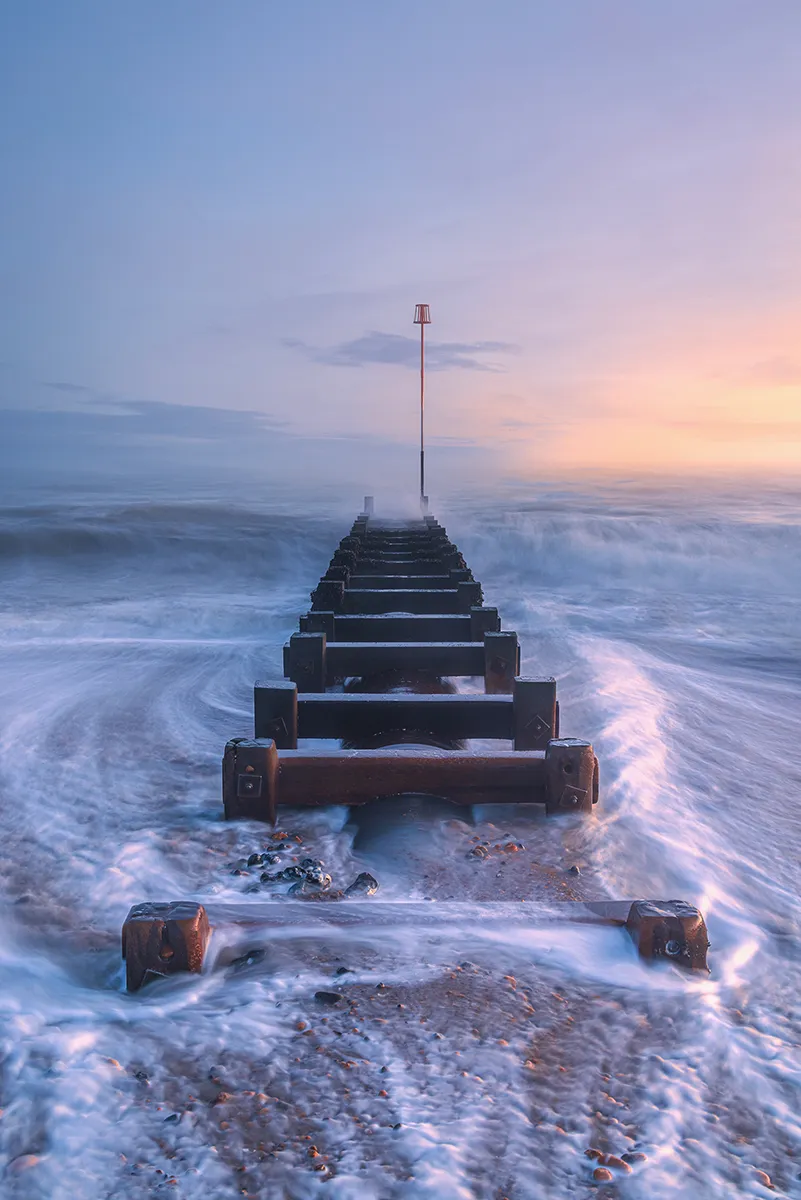 A wooden groyne leading into swirling water beneath a pastel sky in Worthing